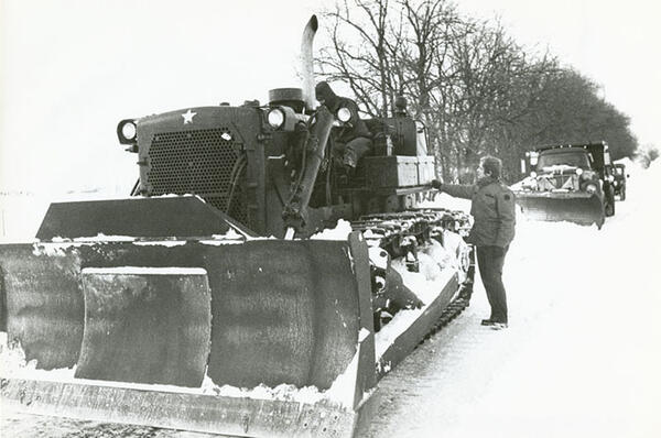 A snow plow with one man behind the wheel and another beside it