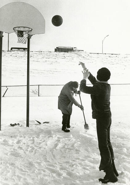 A young boy shooting a basketball into a hoop on a court covered in snow while an adult clears the snow