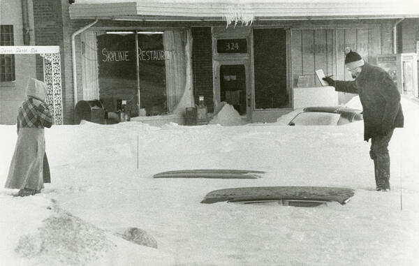 Two people standing on a snowdrift burying their car