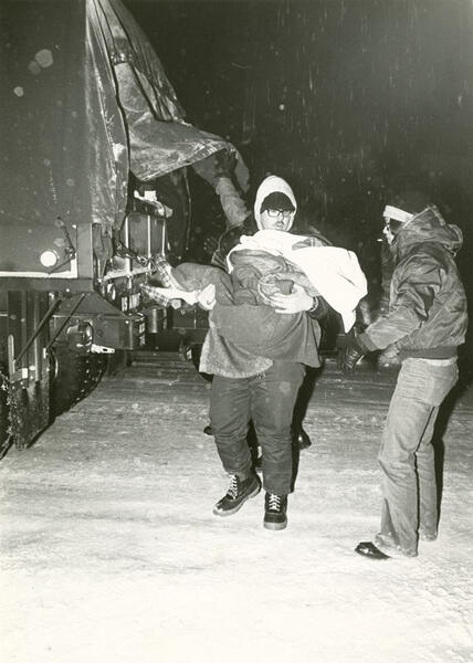 A man carrying a child in a snowstorm with another person assisting