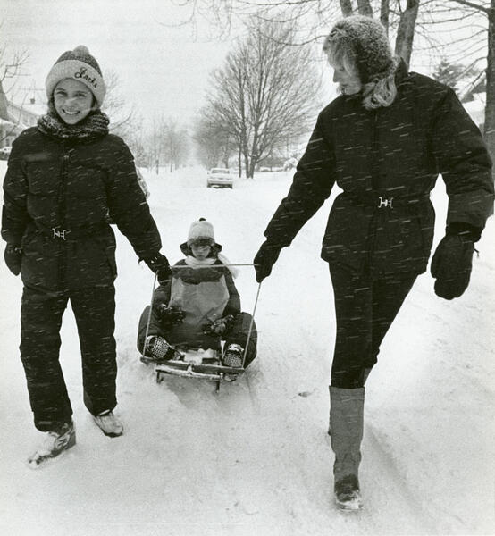 A mother and teen in snow gear pulling a child in a sled down a street in the snow