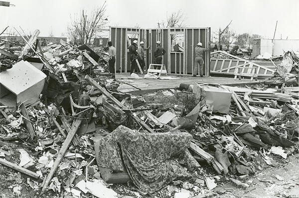 Construction workers raise two walls while surrounded by heaps of tornado debris.