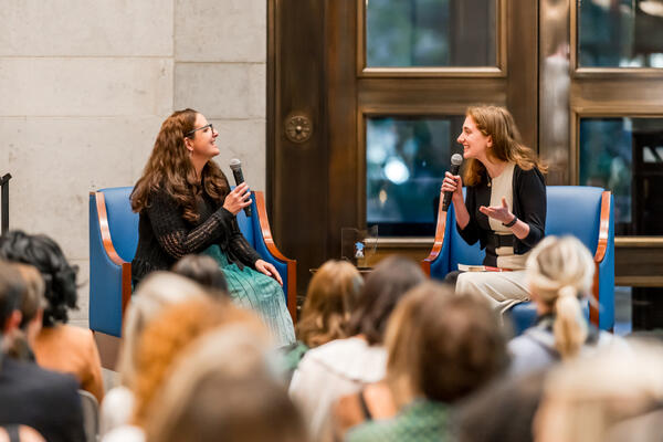 Two people seated on stage facing each other holding microphones in front of an audience.