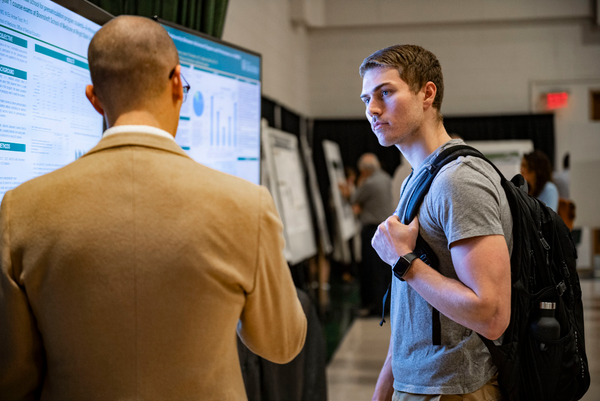 Two men standing in front of a presentation board; one talking and one listening.