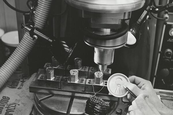 A close-up of a pop-top can end being held by a Dayton Reliable tool employee (only the hand is visible) in front of a machine press. 