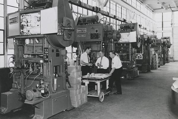 A digitized, historic image depicting six large metal cutting machines on the  manufacturing floor of Dayton Reliable Tool Company in the 1960s. Three men are standing near one of the machines, with Ermal Fraze in the middle.