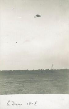 An early aircraft flying over a field above a small crowd and launching derrick.