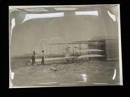 A historic photograph of the Wright Brothers in a field next to their aircraft, in a mylar encapsulation.