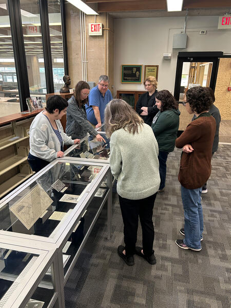 A group of people gathered around an exhibit case. One person is pointing to an item and others are looking in that direction.
