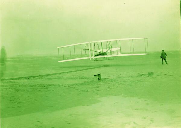 A person flying an early aircraft and another person watching (first flight photograph). The entire photo is green.
