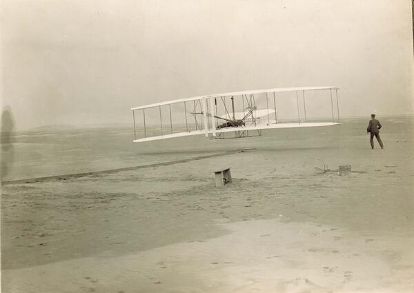 A person flying an early aircraft and another person watching (first flight photograph).