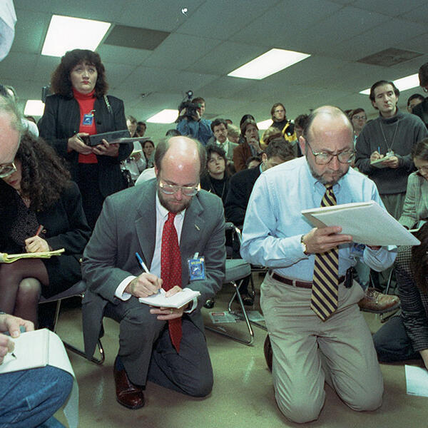 A group of journalists taking notes; three kneeling in front of the camera.