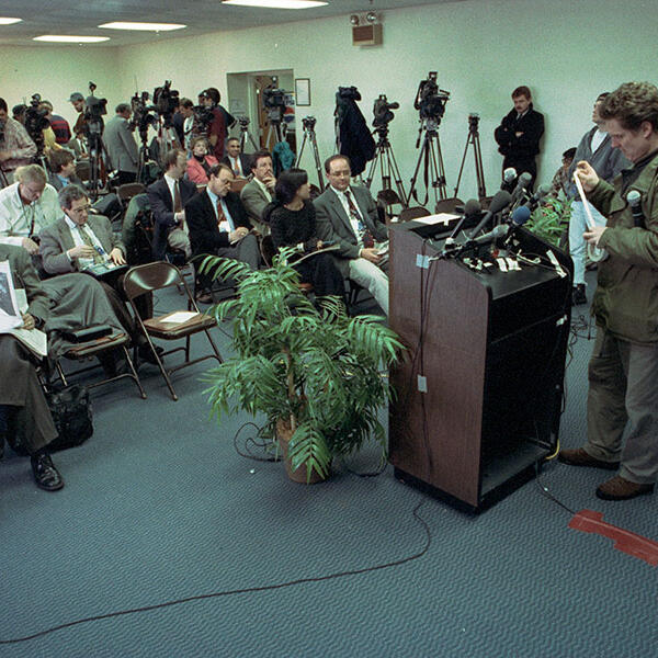 A group of journalists waiting in a room; a podium and camera tripods are visible.