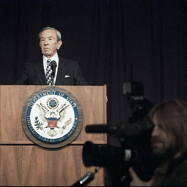 Secretary of State Warren Christopher at a podium with a camera man in the foreground.