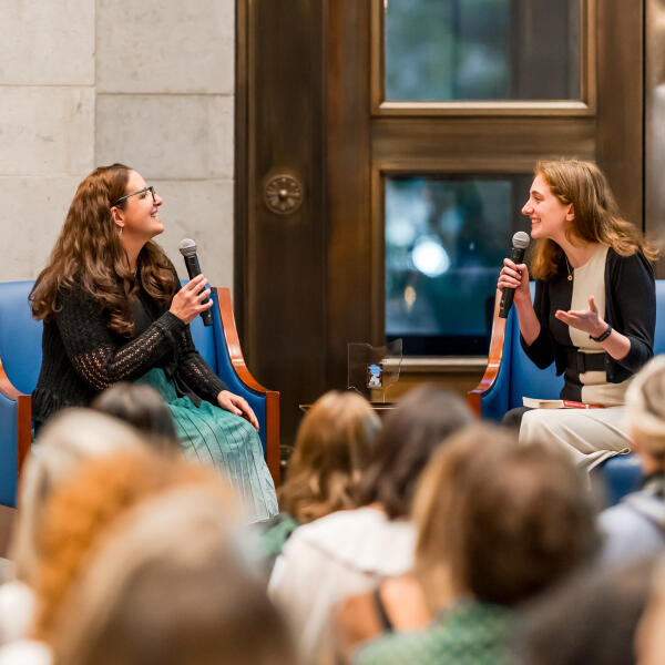 Two people seated on stage facing each other holding microphones in front of an audience.