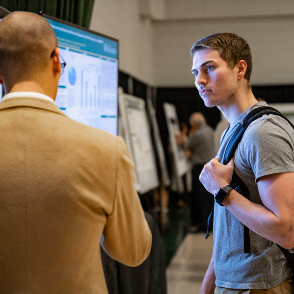 Two men standing in front of a presentation board; one talking and one listening.